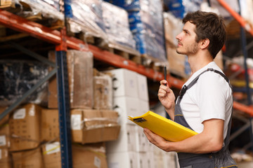 Obraz premium Side view of handsome worker concentrated on goods, standing near shelves in warehouse. Man holding pen and yellow clipboard. Specialist wearing white t shirt and uniform.