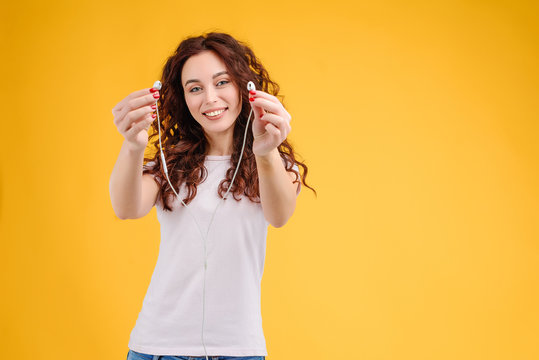 Attractive brunette girl with curly hair isolated over yellow background having fun, listening to music, singing and dancing with earpods on. Music app and online karaoke concept