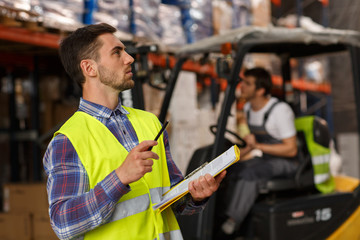 Two workers of warehouse concentrated on their job. Professional manager in reflective waistcoat checking and counting boxes and goods. Forklift driver operating vehicle.