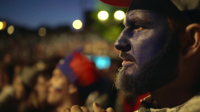 French fan with paint on his face intensely watching football game, experiencing. Man hold his hand in face of excitement sport match. guy worried against background crowd from loss his favorite team