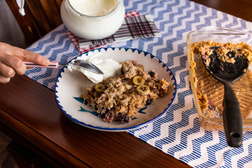 preperation of baked oatmeal with banana, blueberries, walnut and raisins. served with yoghurt and honey. dripping honey. putting yoghurt.