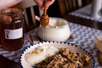preperation of baked oatmeal with banana, blueberries, walnut and raisins. served with yoghurt and honey. dripping honey. putting yoghurt.