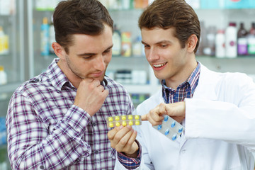 Male pharmacist standing with client in drugstore and helping. Man in white coat holding blister packs with yellow and blue pills. Male customer thinking and holding hand near chin.