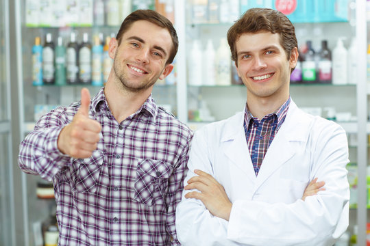 Two good looking men standing in drugstore smiling and posing. Handsome pharmacist in white coat looking at camera and male customer in checked shirt showing thumb up.