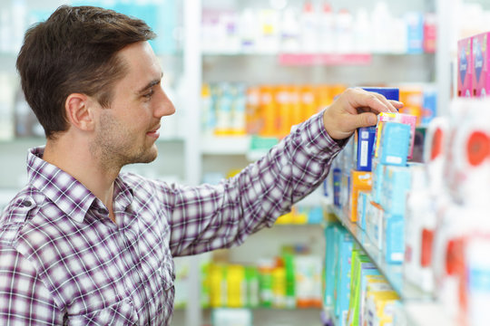 Side view of man wearing in checked shirt choosing medicines in drugstore. Male customer standing, looking at shelf with medical products and taking medicament in hand.