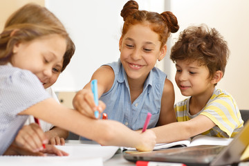 Cheerful schoolgirl sitting at table near friends and drawing with pen on hand of classmate. Happy...