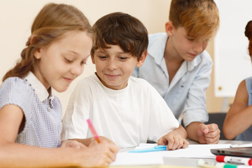 Smiling schoolboy talking with female classmate while leaning new material in classroom. Smart little student sitting near girl and looking at her copybook. Concept of learning and help.