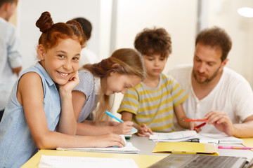 Fototapeta premium Beautiful schoolgirl learning new material during lesson in classroom. Happy cute pupil sitting at table, looking at camera and smiling. Concept of interesting and modern education.