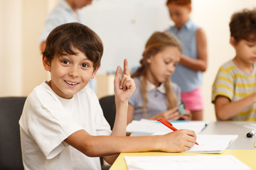 Little surprised boy thinking up idea for writing during lesson in classroom. Inventive student sitting at table, taking notes, looking at camera and holding finger up. Concept of new idea.