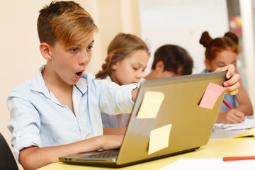 Astonished pupil with open mouth sitting at table and looking at computer in classroom. Stylish boy studying and shocked with new hard material. Concept of education and school.
