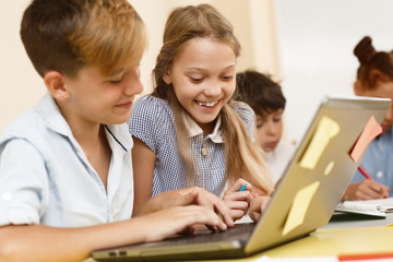 Happy schoolgirl helping classmate in studying new material on computer in сlassroom. Pretty little girl sitting near boy and typing in his computer during lesson. Concept of help and teamwork. - obrazy, fototapety, plakaty