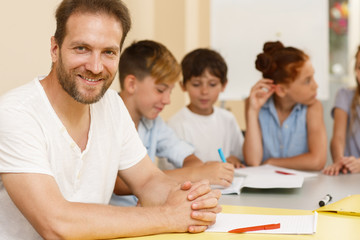 Intelligent teacher sitting at table, looking at camera and posing in school. Qualified tutor teaching little students, giving them tasks and problems in classroom. Concept of interesting education.
