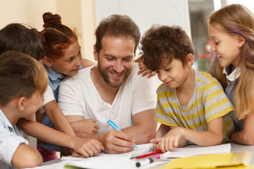 Fototapeta premium Tutor sitting at table, writing and explaining material, smiling children standing around and listening. Happy male teacher in process of teaching little students in classroom. Concept of education.