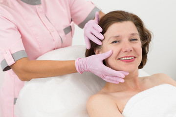 Smiling woman lying on couch during anti aging procedure  and enjoying facial massage in cosmetology office. Hands of cosmetologist touching skin of face. Concept of beauty and care.