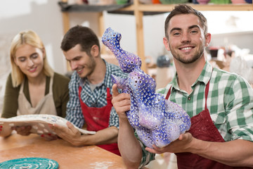 Positivity craftsman posing near table keeping blue product of clay and colleague standing back. Three masters in aprons admiring unusual work of art in workshop. Concept of teamwork.