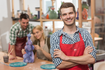 Portrait of handsome craftsman in checkered shirt and red apron wit folded arms looking at camera, smiling and posing during pottery class. Cheerful man enjoying new favorite hobby in art studio.