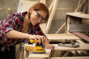 Fototapeta na wymiar Professional and talented craftswoman smiling and working with electric sender and lumber in carpentry shop. Beautiful and positive woman wearing checked shirt and safety glasses.