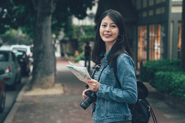 young chinese woman tourist standing in street with backpack hold map geography. female travel photographer visit carmel by the sea old town country in america face camera smile. happy girl by road
