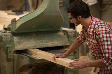 Professional and busy joiner working with wood and carpenter's gauge in joiner's shop. Confident woodworker in checked shirt standing, holding timber and machining it.