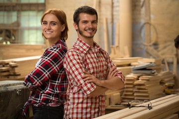 Pretty craftswoman with ginger hair and handsome craftsman posing in joiner's shop. Bearded joiner and beautiful woman smiling and looking at camera. Team wearing in checked shirts.