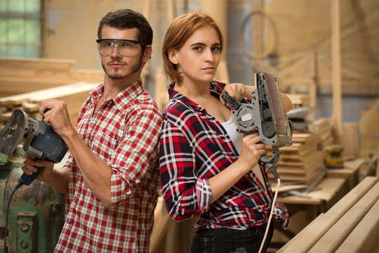 Male and female joiners holding grinding machines for machining wood. Handsome bearded man in safety glasses and beautiful woman with ginger hair looking at camera and posing.