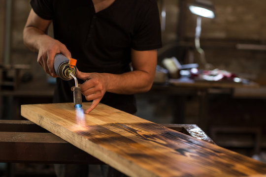 Crop of man wearing in black shirt, burning wooden board for displaying textures with petrol blowtorch ignition at factory. Male employee board carefully for producing best pattern.