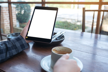 Mockup image of woman's hands holding black tablet pc with blank screen while drinking coffee on wooden table in cafe