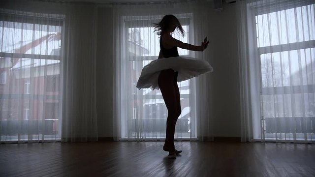 A studio in twilight. A silhouette of young woman ballerina performing a spin in front of the window