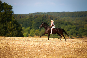 Horse with rider (woman) on a summer evening gallops over a harvested cornfield, wearing only a summer dress and bareback with open hair.