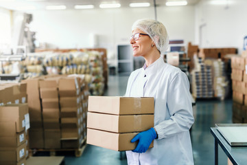 Smiling blonde employee in sterile uniform and with eyeglasses holding boxes while standing in food factory.