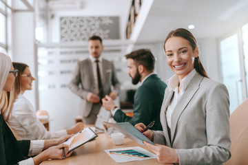 Gorgeous smiling businesswoman in formal wear and long brown hair writing down analyze of business success and looking at camera while sitting at meeting in board room. Colleagues listening to CEO.