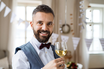 A portrait of a mature man standing indoors in a room set for a party, holding wine.