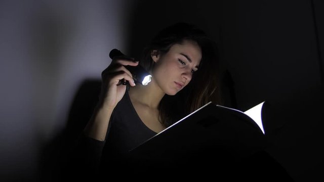 close-up portrait girl is reading a book with a flashlight in a dark room at night. 4k. Motion shooting