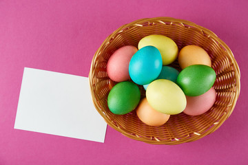 Beautiful easter table composition. Pile of painted eggs of different passtel colors in wicker basket, blank page on crimson paper background. Greeting card template. Copy space, close up