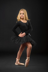 Fototapeta premium Cute little ballerina with long hair in a black tutu and pointe posing on a black background