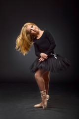 Cute little ballerina with long hair in a black tutu and pointe posing on a black background