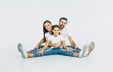 Fototapeta na wymiar Relationship concept. Beautiful and happy smiling young family in white T-shirts are hugging and have a fun time together while sitting on the floor and looking on camera.