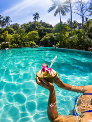 Cropped shot of girl holding coconut cocktail in outdoor swimming pool. Sunny day in tropical...