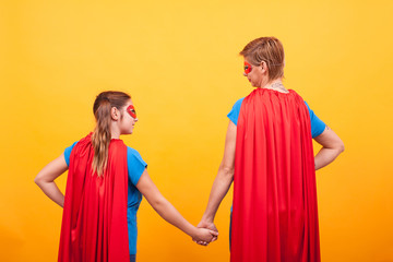 Back view of mother and her little girl dressed like superheros holding hands over yellow background .