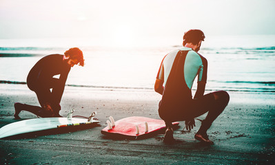Young surfers preparing the gear equipment on the beach at sunset - Main focus on right man body
