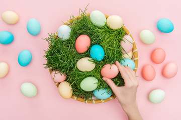 Fototapeta na wymiar top view of female hand with easter eggs in wicker plate with grass isolated on pink