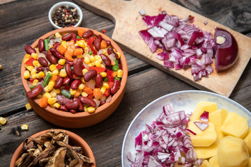 Mixture of vegetables, dried boletus mushrooms, sliced potato, red onion sliced and whole on a cutting board and spices, on old rustic wooden table.