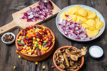Mixture of vegetables, dried boletus mushrooms, sliced potato, red onion sliced and whole on a cutting board and spices, on old rustic wooden table.