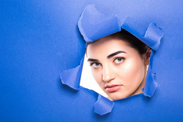 the face of a young beautiful girl with a bright make-up and puffy blue lips peers into a hole in...