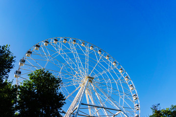 A new very tall white Ferris wheel with comfortable booths for people is slowly rotating in a circle in the center of a big city against a clear blue sky