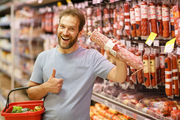 Front view of happy customer holding salami in hand and showing thumb up with another hand. Handsome man smiling, posing and standing in meat section of supermarket.