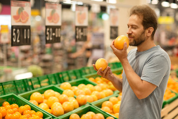 Handsome man holding grapefruits in hands and smelling. Happy facial expression with closed eyes of customer. Bearded man do shopping in supermarket and choosing fresh fruit.