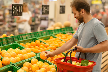 Male customer holding fresh orange and choosing citruses. Bearded man standing in supermarket in fruit section and do shopping. Man holding red basket full of products.