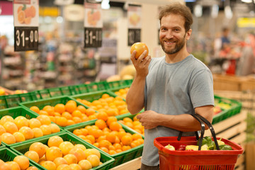 Handsome man posing, looking at camera and holding orange in hand. Bearded customer smiling and holding red basket full of products. Section with fresh citruses on background.
