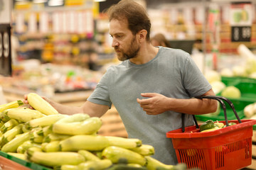 Pile of zucchini and man choosing vegetables in supermarket. Handsome buyer do shopping in vegetable section. Bearded man holding red basket on hand. Concept of gastronomy.